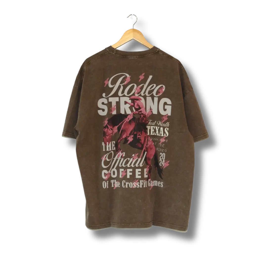 A brown t-shirt with text and graphic prints, hanging on a hanger. The text includes 'Rodeo Strong' and 'The Official Coffee of The CrossFit Games'.